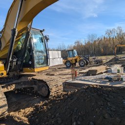 FAVOR - ANDRII SKLIAR - Żółty koparko-ładowarka CAT na placu budowy, w tle zaplecze budowlane, materiały budowlane i maszyny. Słoneczny dzień, błękitne niebo.