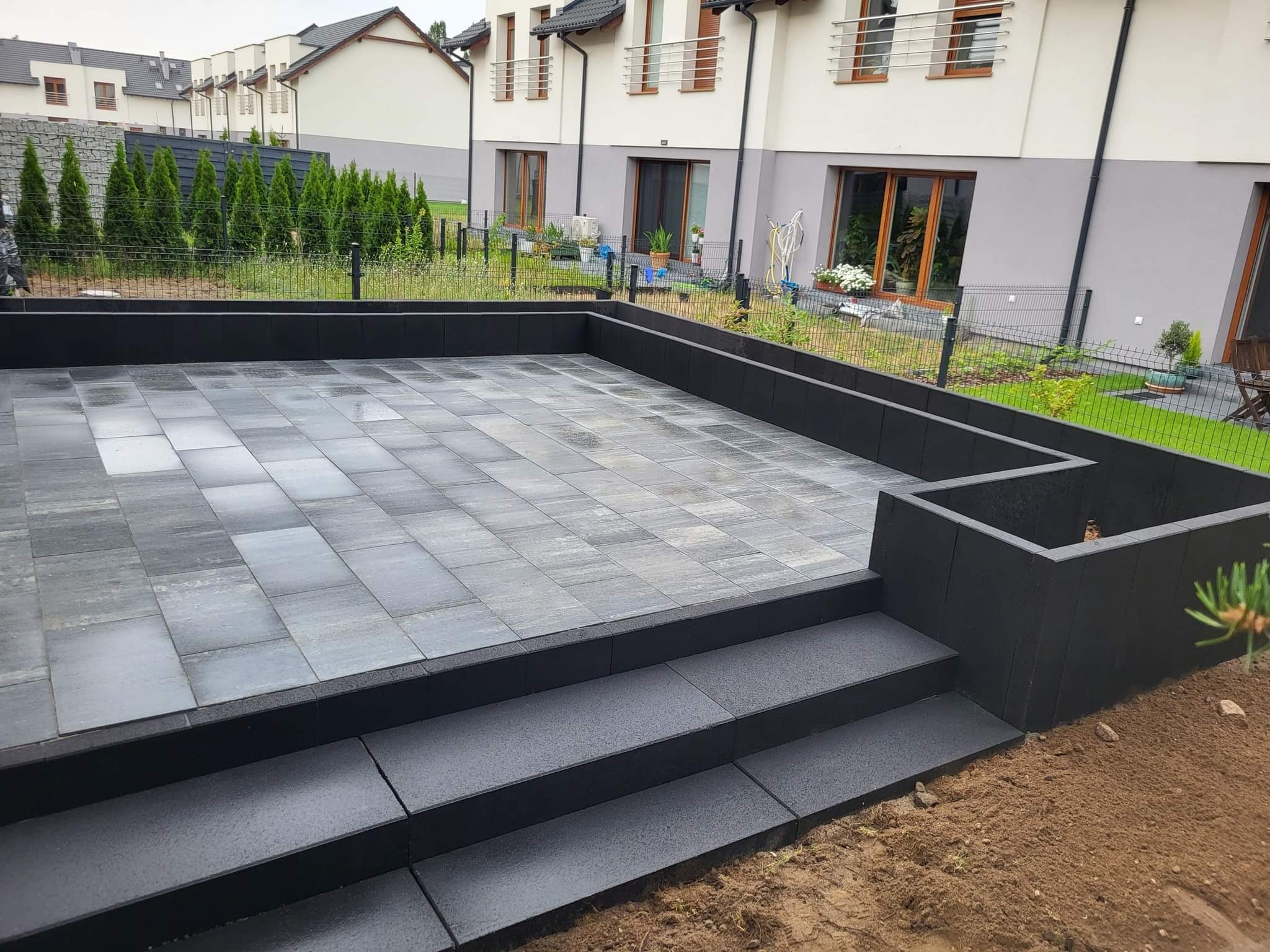 Nowoczesny taras z szarej kostki brukowej, otoczony niskim, czarnym murkiem. Widoczne schody z kostki i fragment ogrodu z ogrodzeniem panelowym.