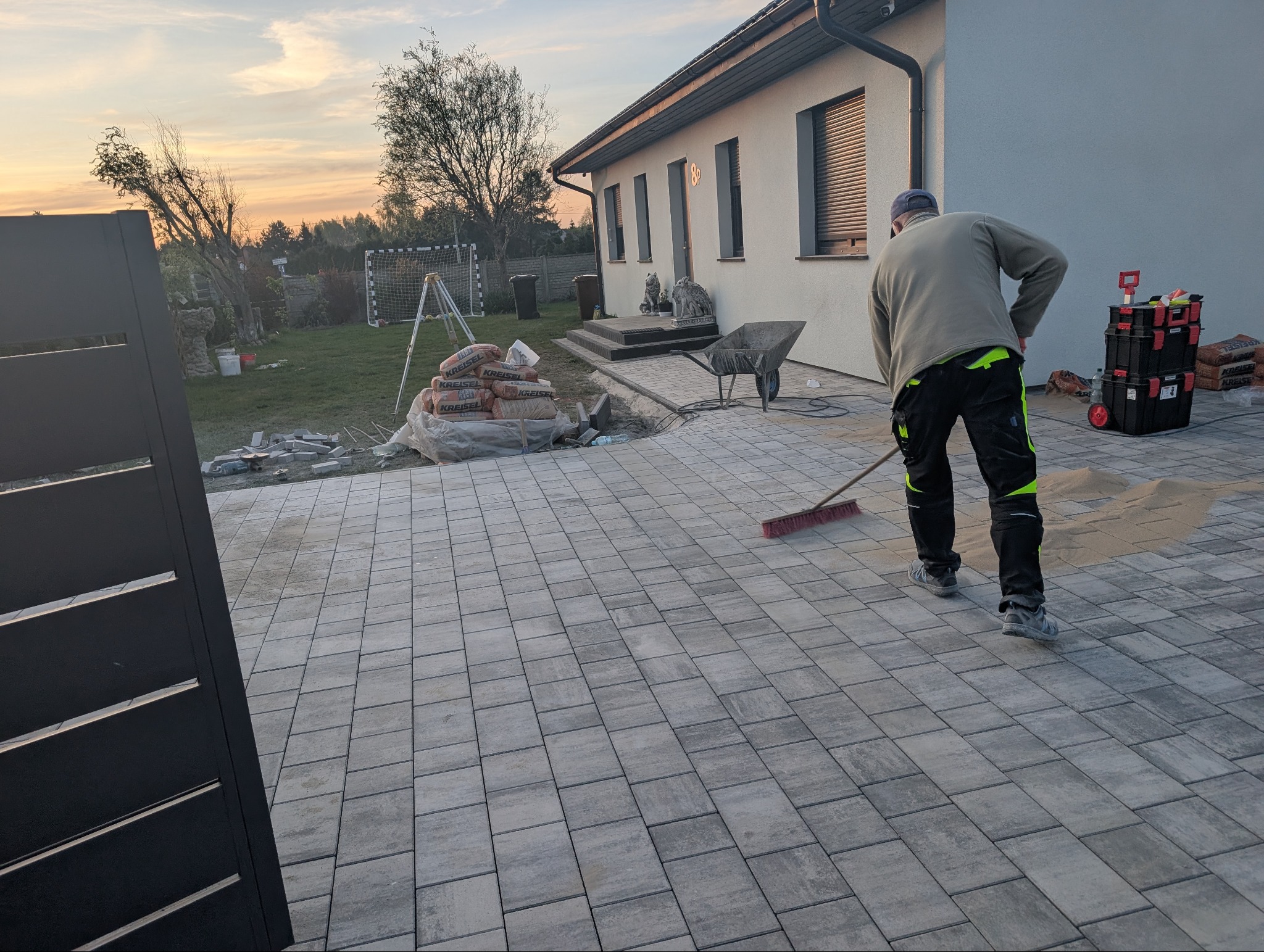 Pracownik zamiata piasek na nowo ułożonej, szarej kostce brukowej na podjeździe. Widoczny fragment ogrodzenia, domu i narzędzia pracy.