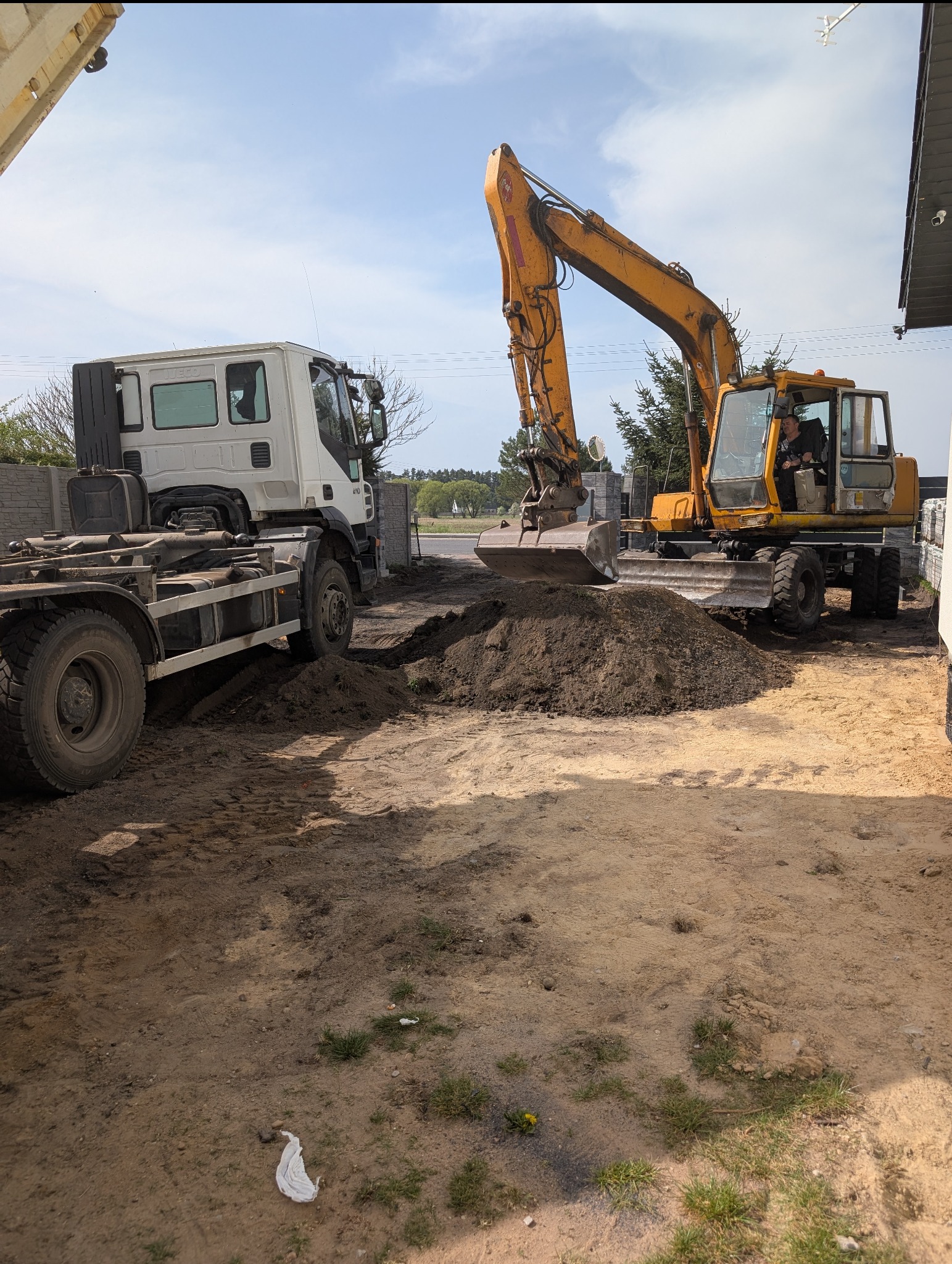 Żółta koparka nabiera ziemię obok ciężarówki na placu budowy. W tle widoczne ogrodzenie i zieleń. Prace ziemne w toku.
