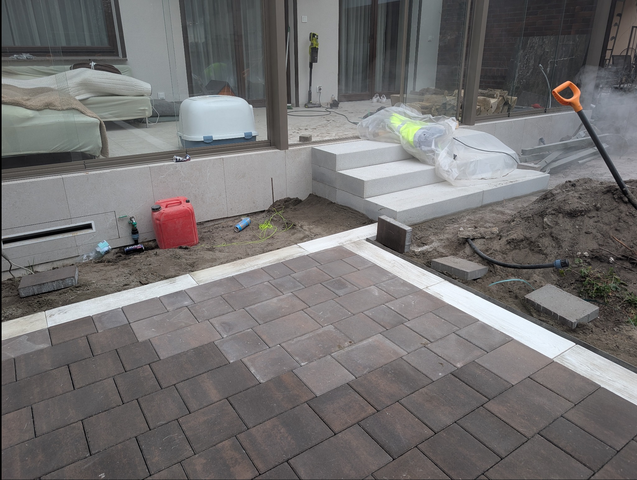 Układanie brązowej kostki brukowej na posesji, widoczne białe obrzeża i narzędzia pracy. Częściowo ułożona nawierzchnia, prace w toku.