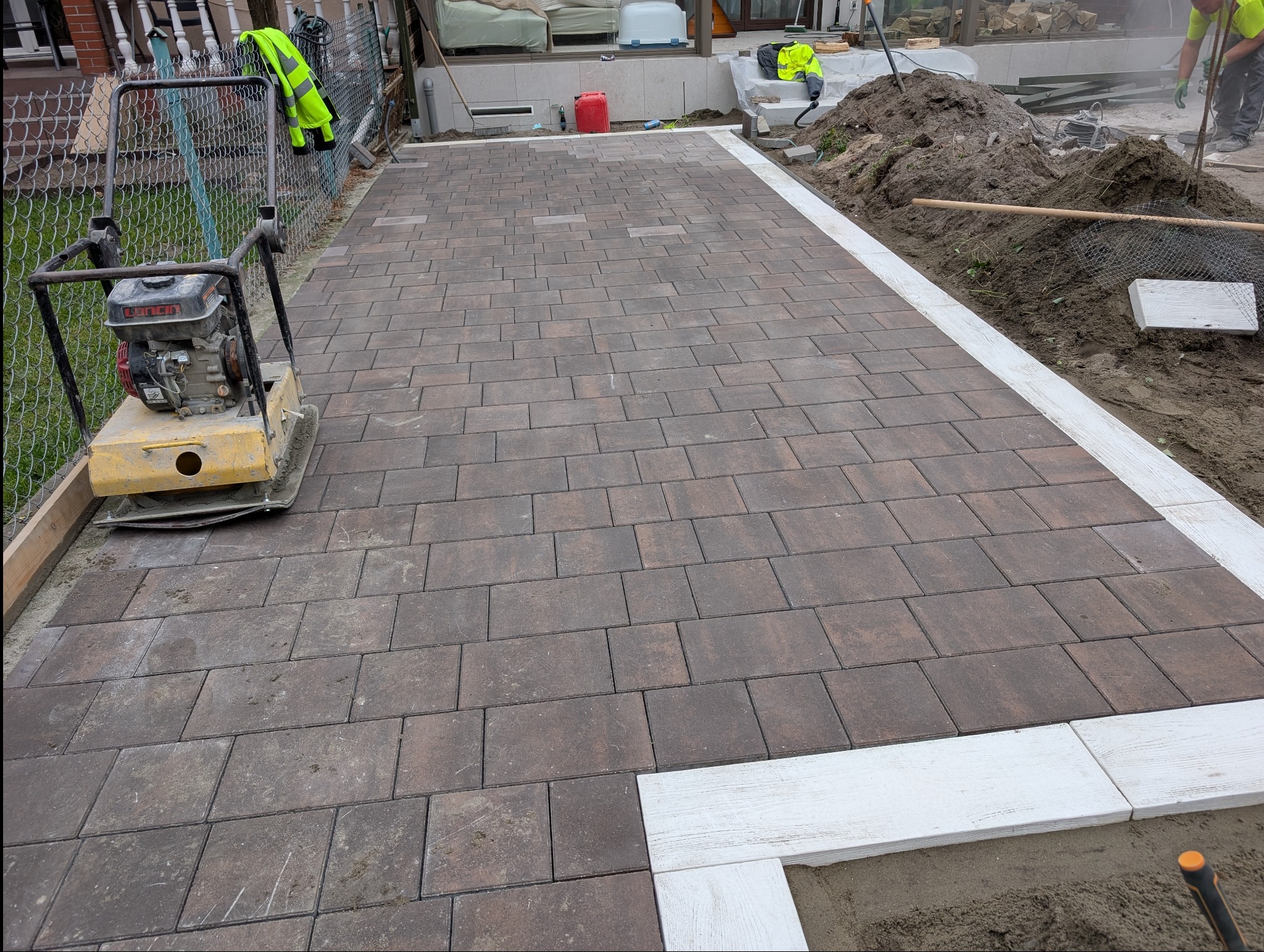 Ułożona, brązowa kostka brukowa z białym obramowaniem. Na pierwszym planie ubijarka. W tle widoczny teren przygotowany pod dalsze prace.