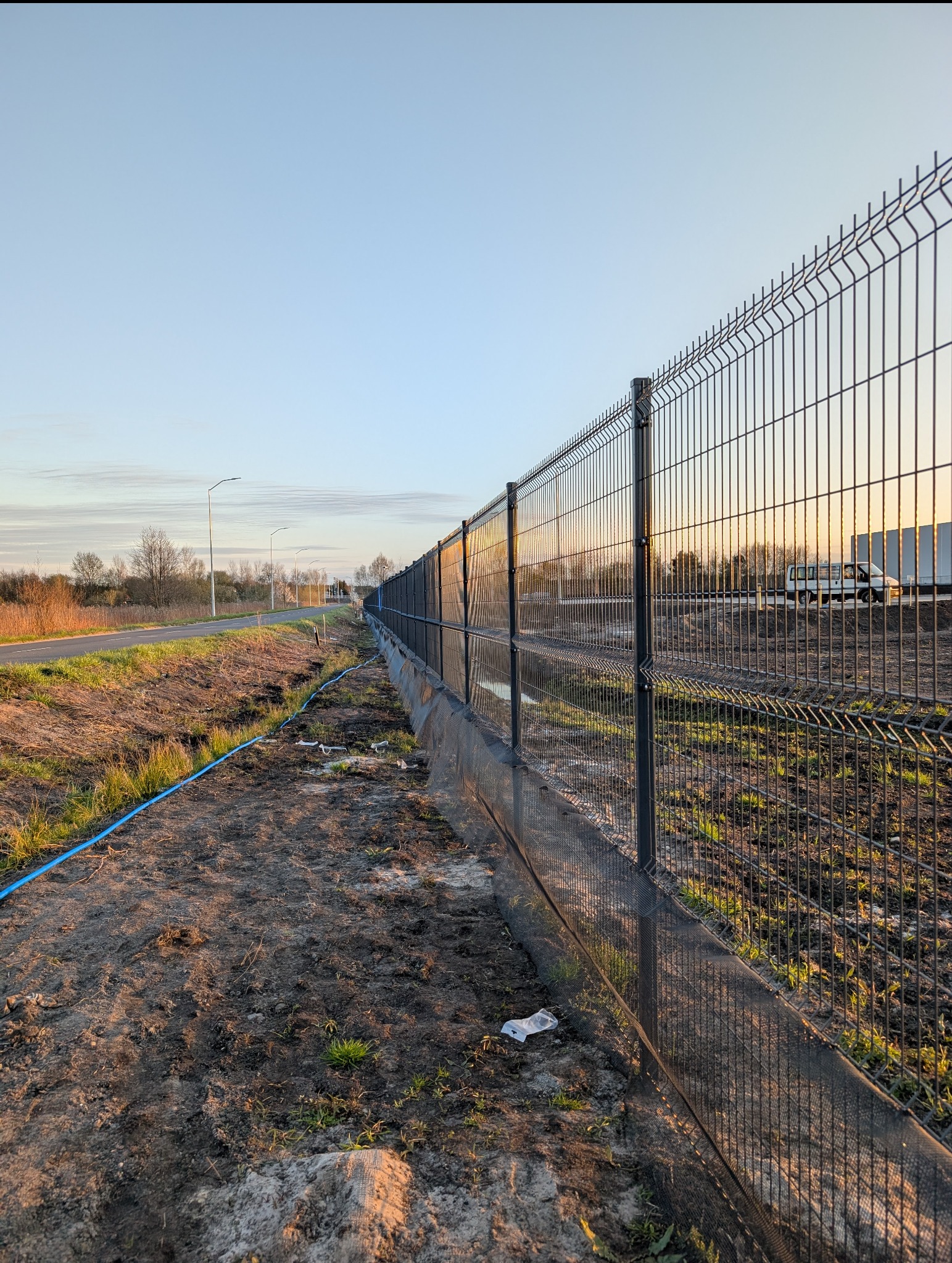 Długi, panelowy, metalowy płot z siatki, zamontowany wzdłuż drogi. Widoczny fragment drogi i pobocze z niebieskim wężem. W tle budynki i pojazd.