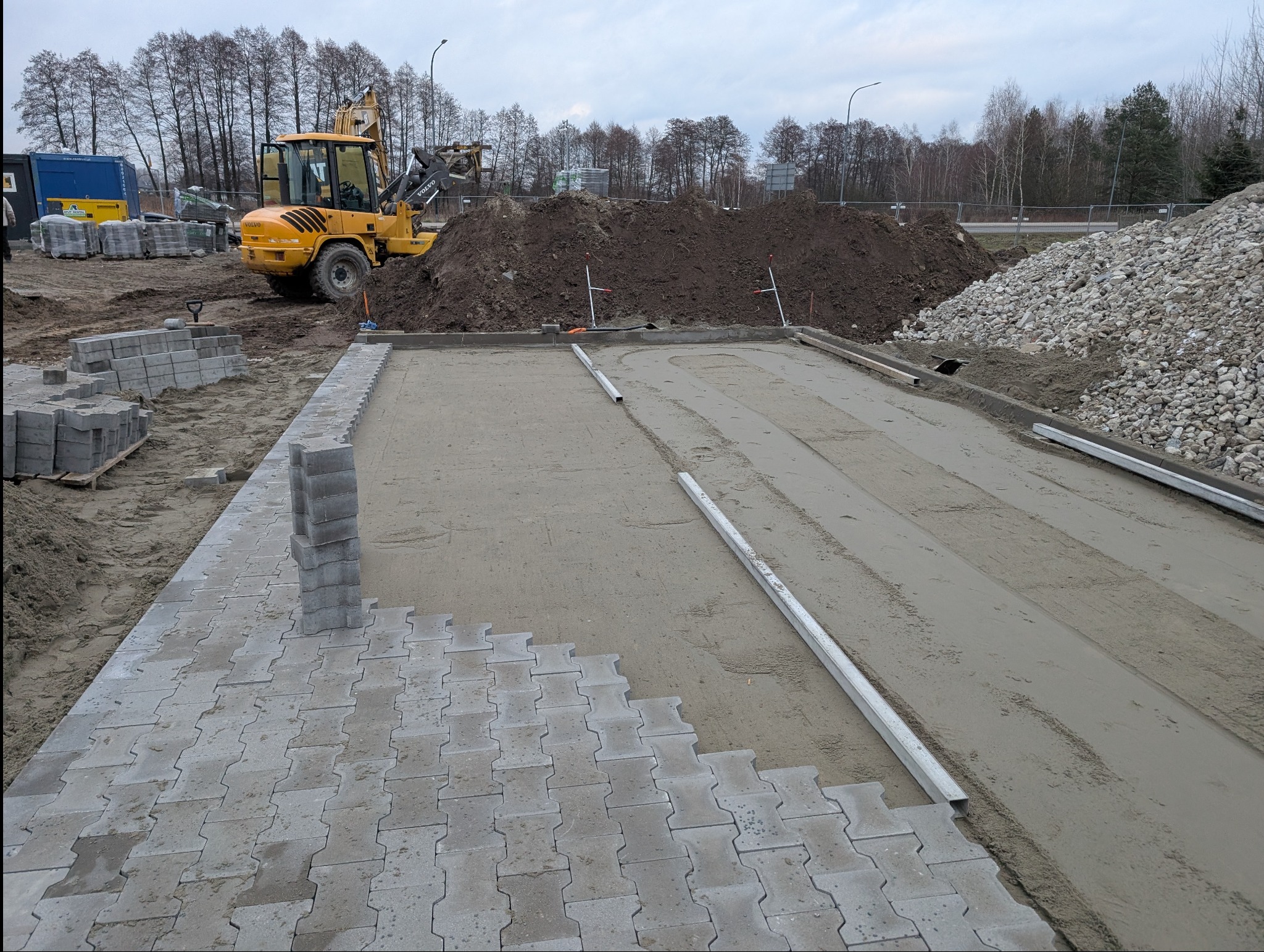 Układanie szarej kostki brukowej na przygotowanym podłożu. W tle żółta koparka i pryzma ziemi. Widoczne narzędzia i materiały budowlane.