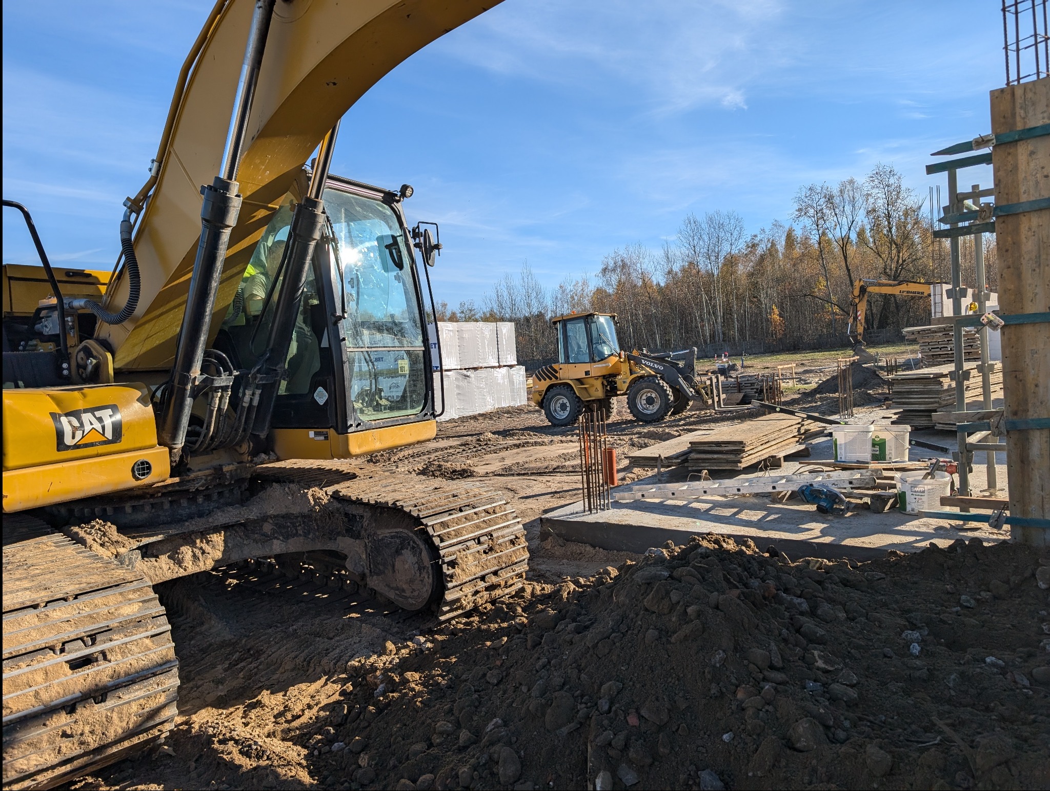 Żółty koparko-ładowarka CAT na placu budowy, w tle zaplecze budowlane, materiały budowlane i maszyny. Słoneczny dzień, błękitne niebo.