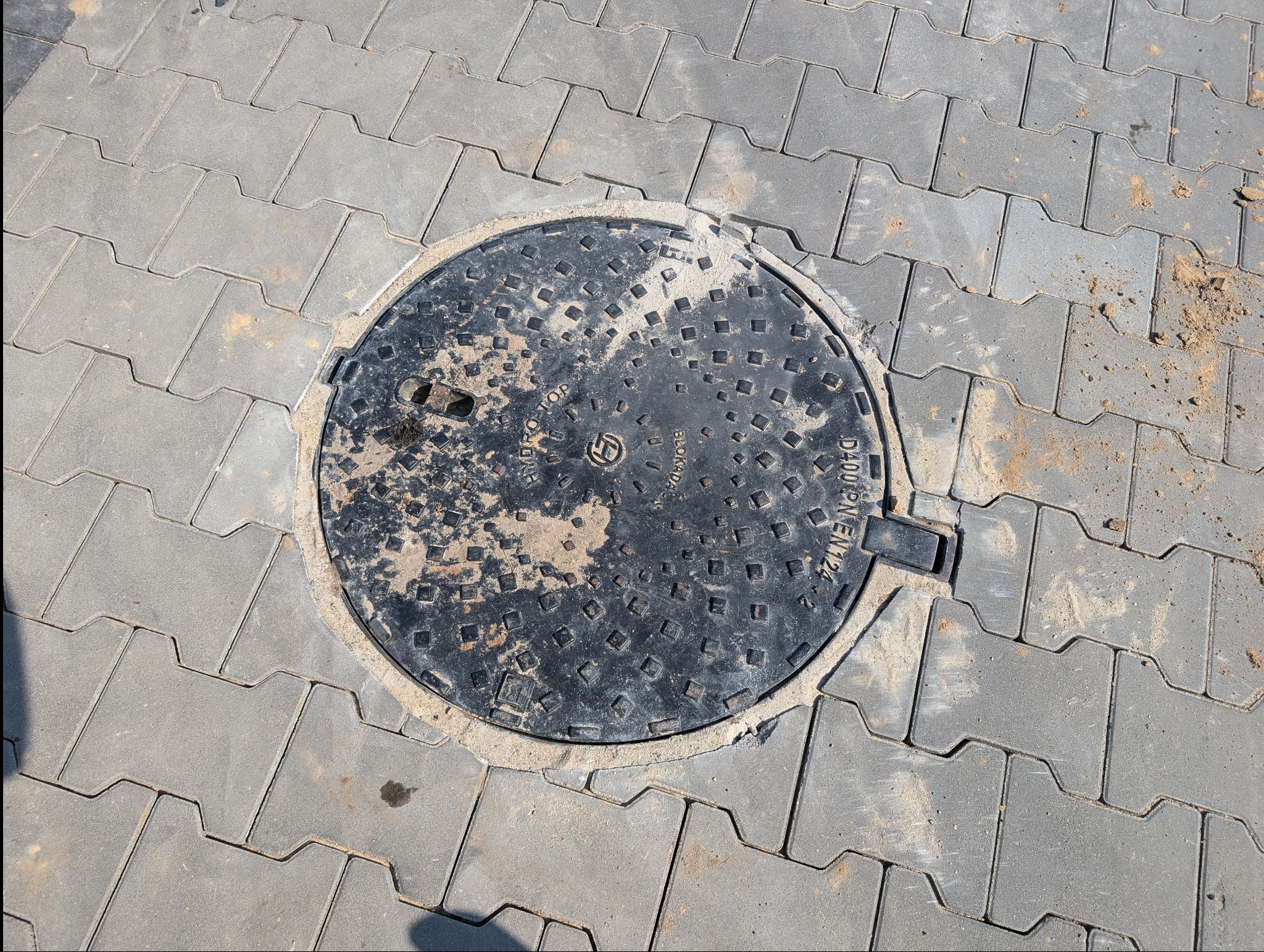 Szara kostka brukowa ułożona wokół czarnej, okrągłej studzienki kanalizacyjnej z metalowym uchwytem, piasek w spoinach.