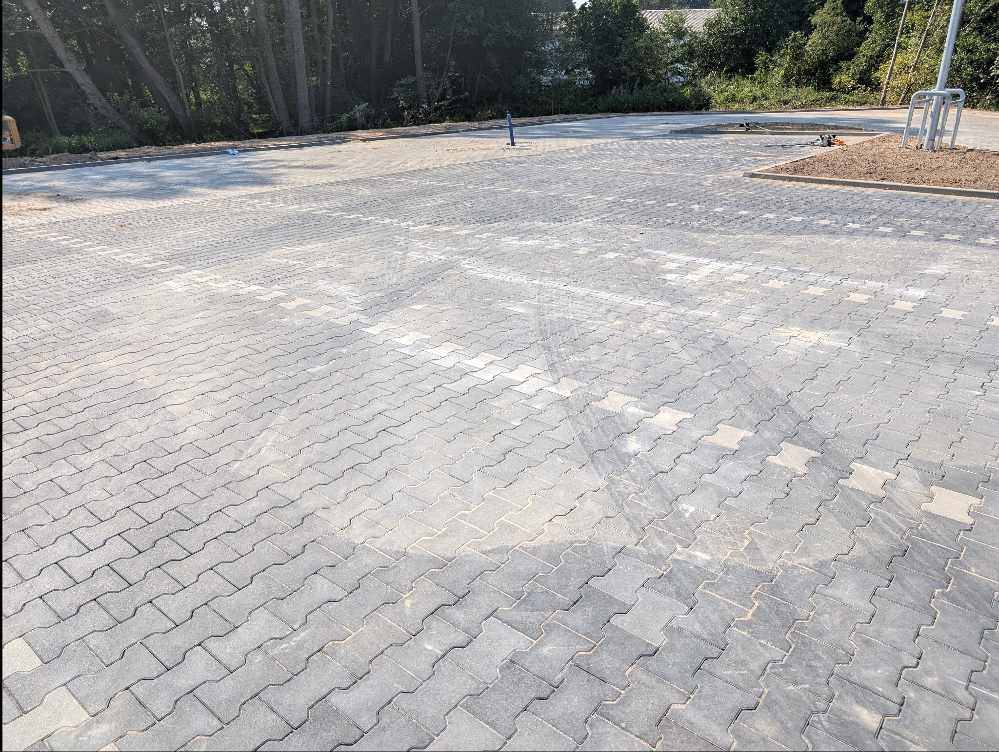 Szara kostka brukowa ułożona w jodełkę na parkingu, z widocznymi śladami po pojazdach. W tle drzewa i elementy infrastruktury parkingowej.