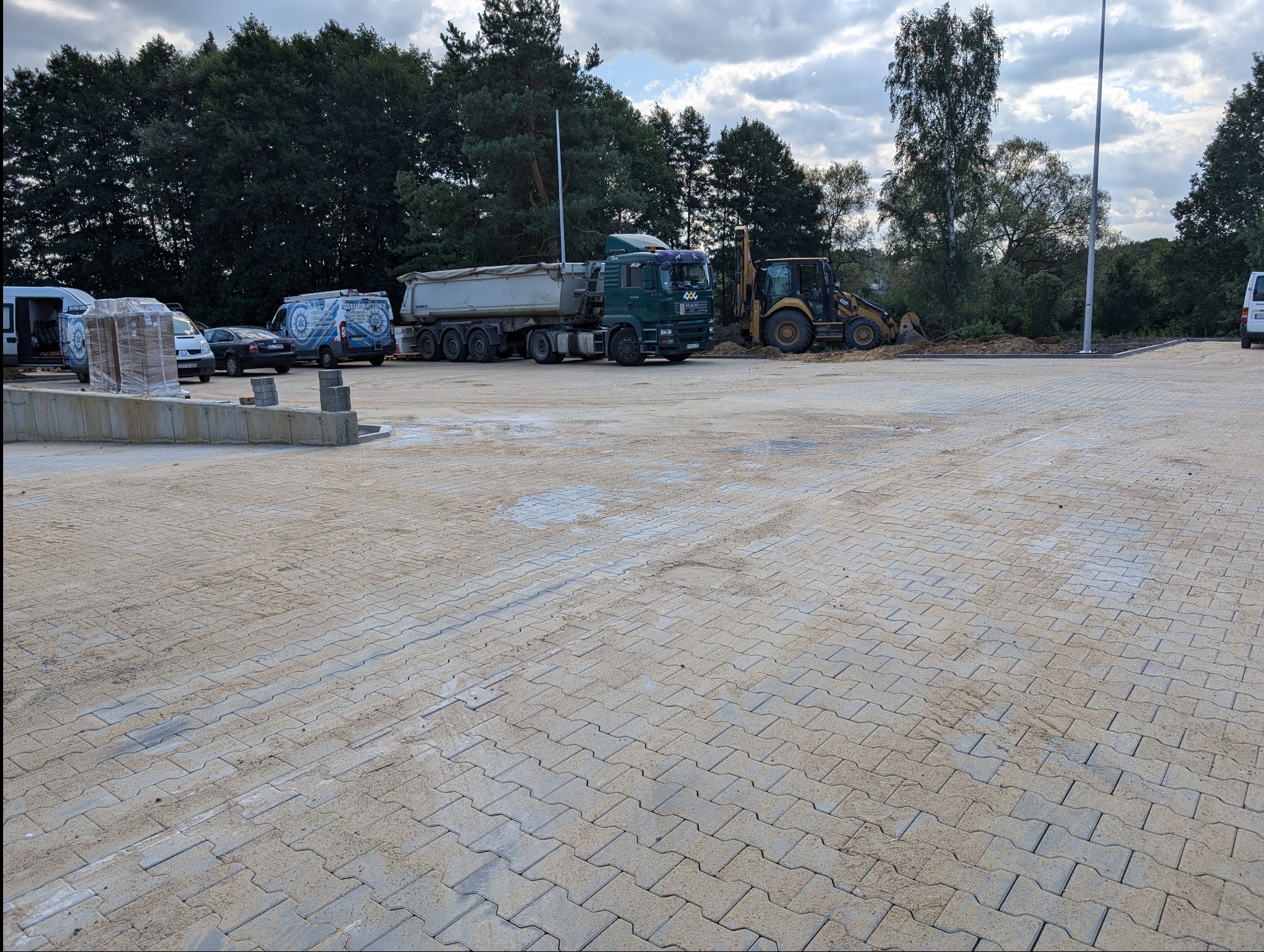 Rozległy plac wyłożony szarą kostką brukową, w tle zaparkowane pojazdy budowlane i samochody firmowe. Widoczne drzewa i pochmurne niebo.