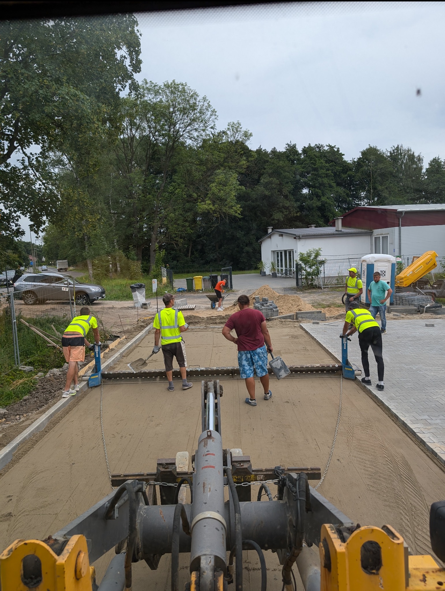 Ekipa w odblaskowych kamizelkach wyrównuje piasek pod kostkę brukową za pomocą specjalistycznej maszyny; widok z kabiny operatora.