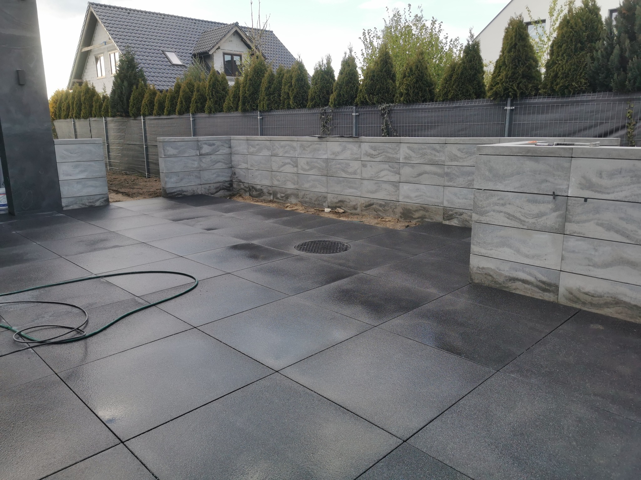 Nowoczesna aranżacja przestrzeni z ciemną kostką brukową, ogrodzeniem z siatki i elementami dekoracyjnymi z szarej kostki. Widoczny fragment węża ogrodowego.