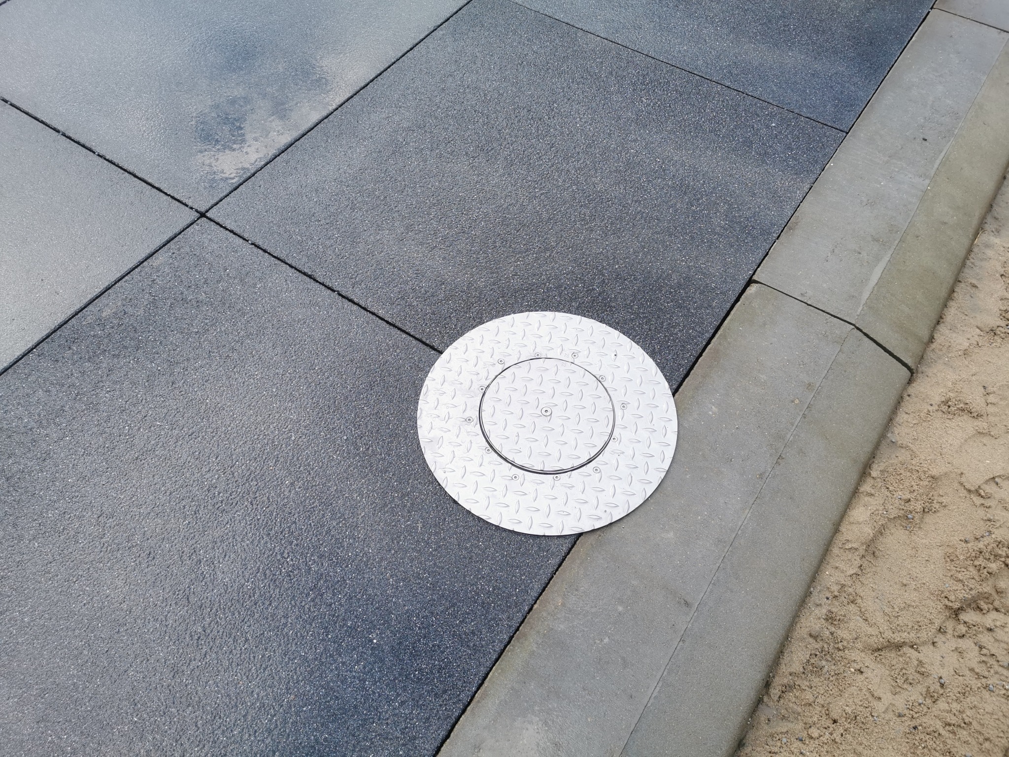 Szara, gumowa kostka brukowa ułożona równo z metalową studzienką rewizyjną i betonowym obrzeżem. Widok z góry na fragment chodnika.