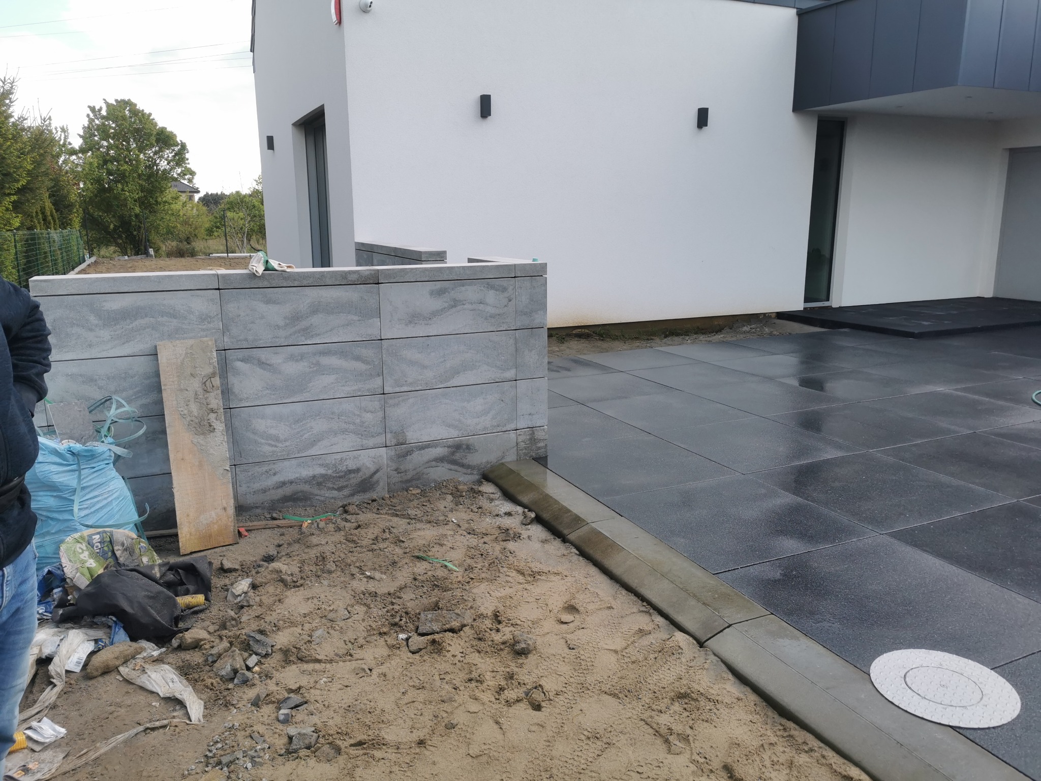 Nowoczesna posesja z szarej kostki brukowej w trakcie układania, widoczne materiały budowlane i fragment budynku z oświetleniem zewnętrznym.