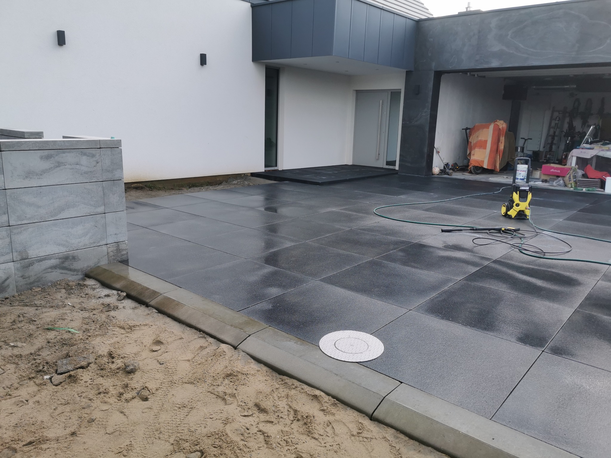 Nowoczesny podjazd z ciemnej kostki brukowej, obrzeża z kostki, widoczny fragment domu z garażem. Częściowo rozłożona kostka brukowa w piasku.