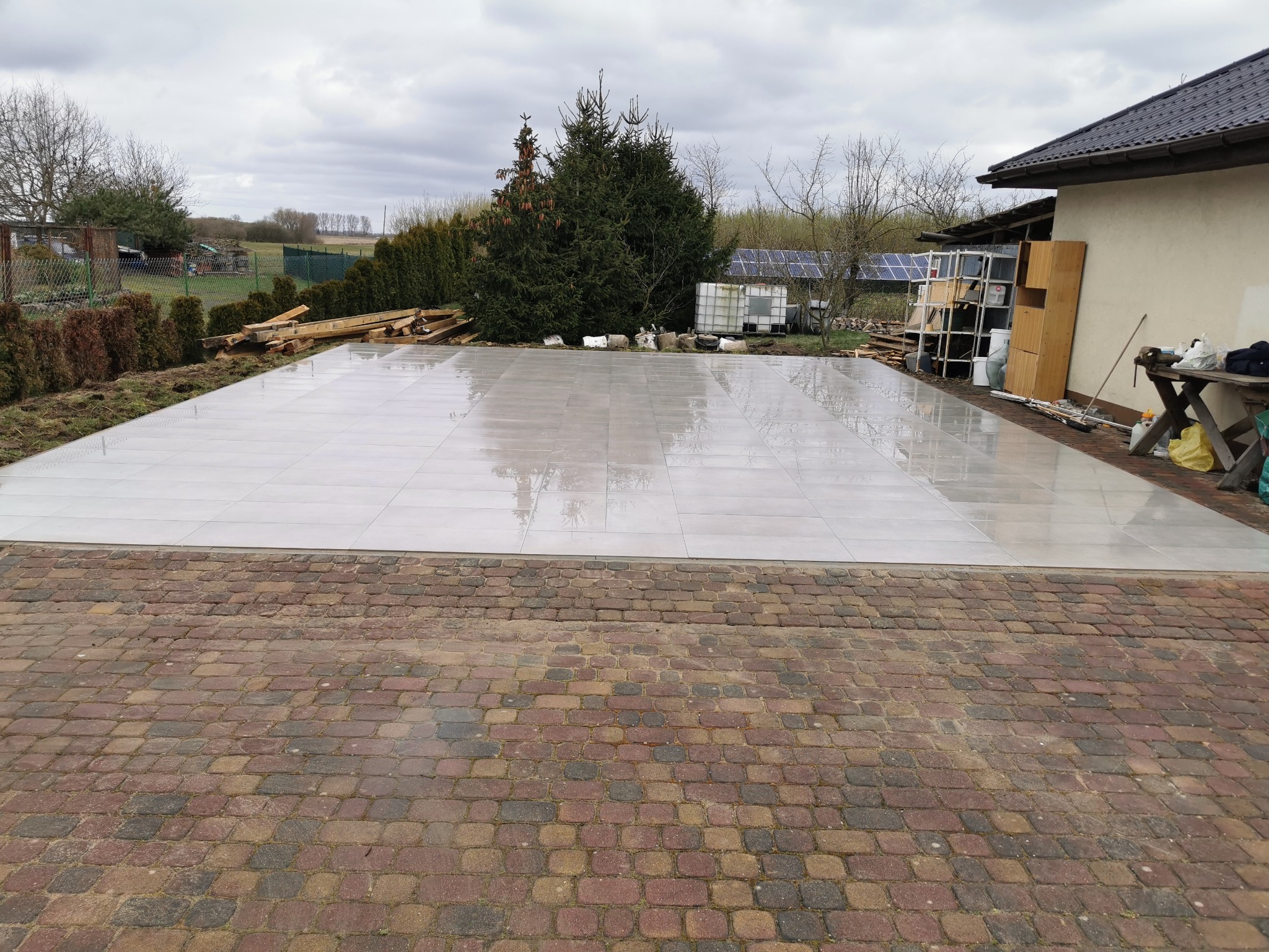 Ułożona, jasnoszara, duża płyta tarasowa na zewnątrz, obok kostka brukowa, widoczne drzewa i budynek w tle. Realizacja z dbałością o detale.