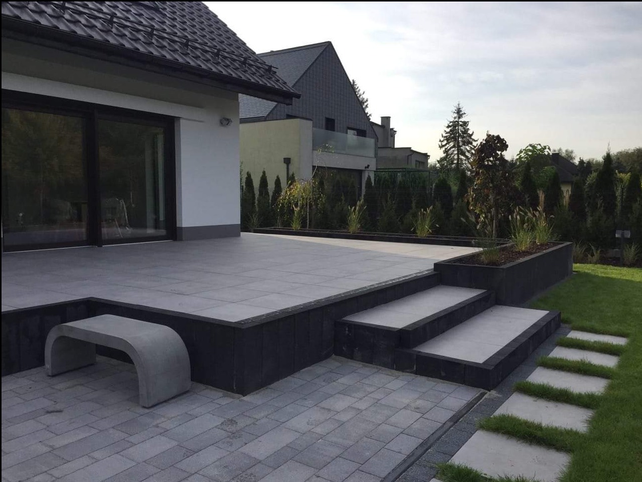 Nowoczesny taras z szarych płyt, schodów i kostki brukowej. Minimalistyczna ławka, donica z roślinami i ścieżka w trawie. Dom z dużym oknem przesuwnym w tle.