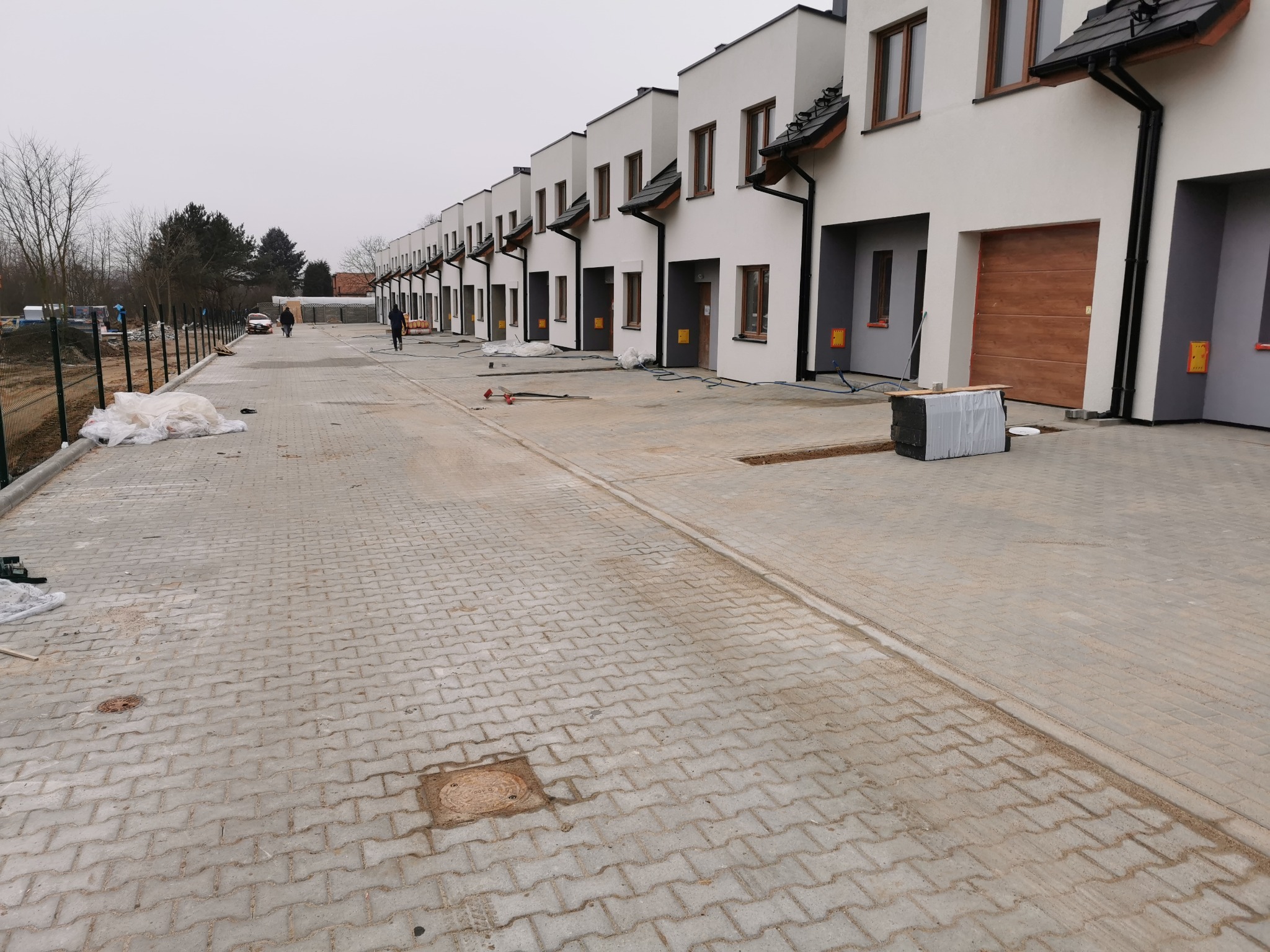 Nowe osiedle domów szeregowych z wybrukowaną drogą dojazdową. Kostka brukowa ułożona w zygzak. Widoczne studzienki kanalizacyjne i przygotowane materiały budowlane.