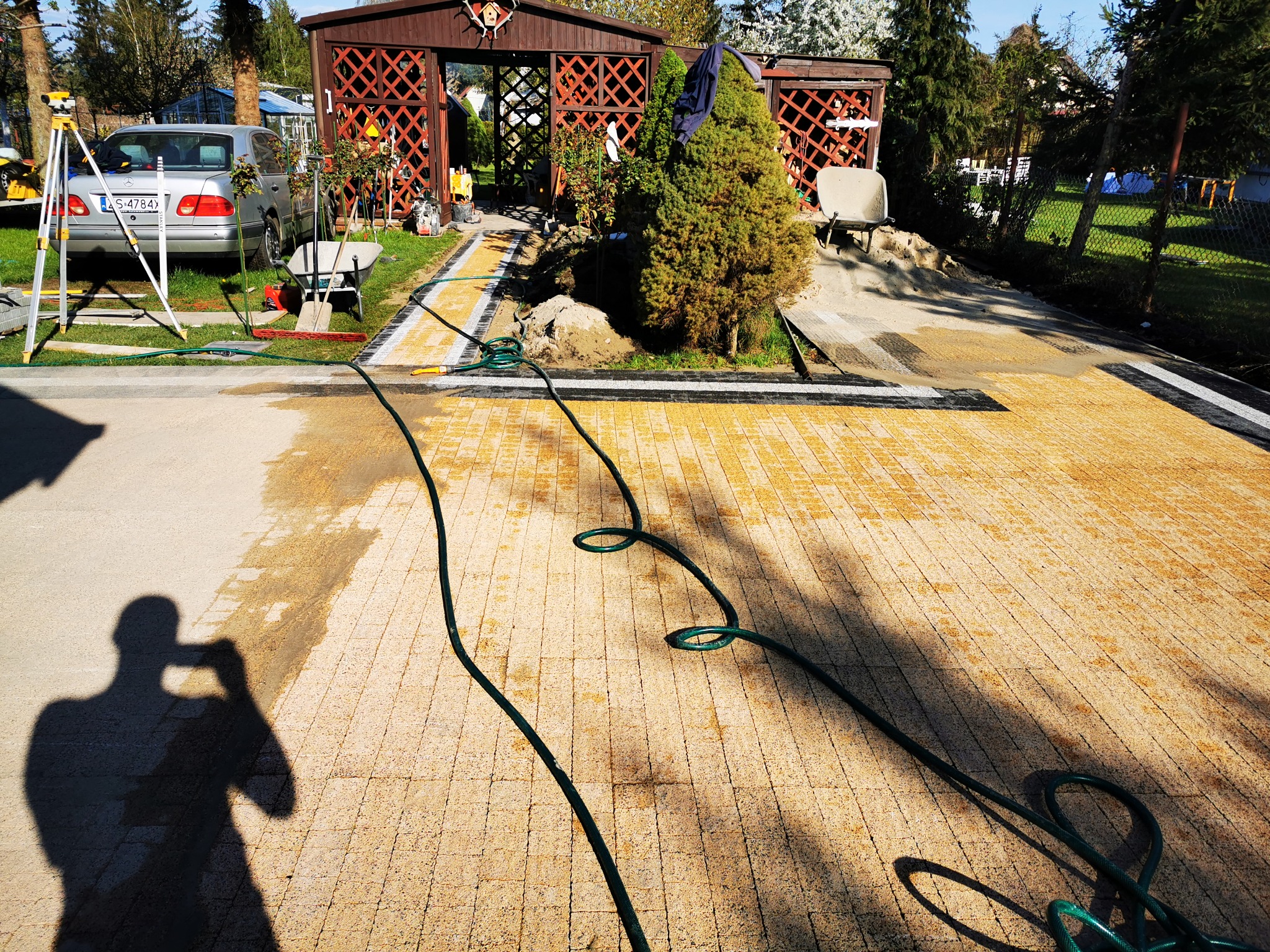 Żółta kostka brukowa ułożona na posesji, widoczny wąż ogrodowy, cień osoby robiącej zdjęcie, samochód, narzędzia i altana w tle. Prace brukarskie w toku.