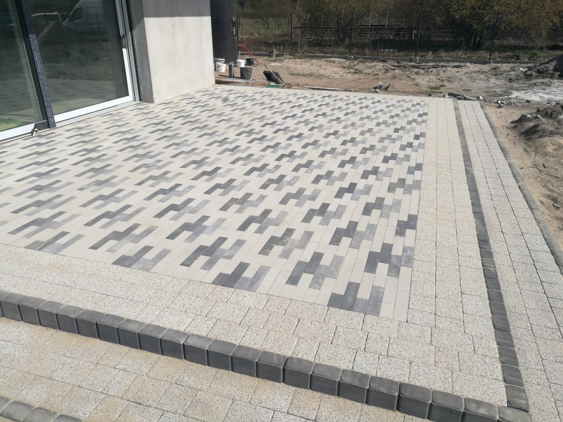 Nowoczesny taras z kostki brukowej w odcieniach szarości i beżu, ułożonej we wzór jodełki. Widoczne detale wykończenia i otoczenie w trakcie prac.