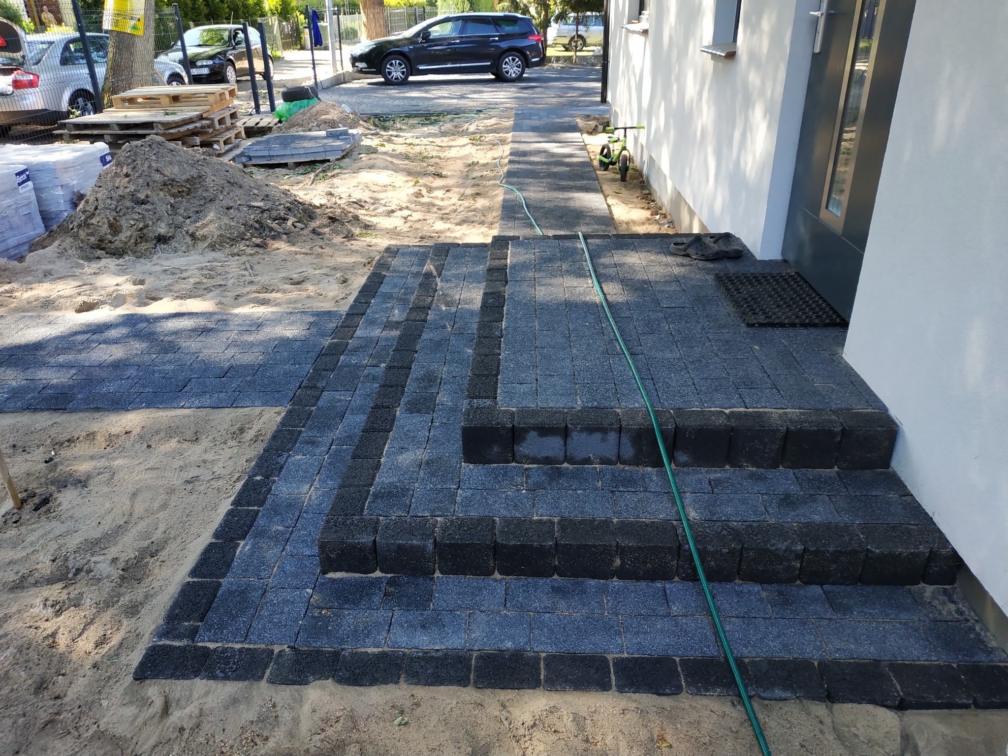 Ułożona kostka brukowa w odcieniach szarości prowadząca do wejścia domu. Widoczny fragment trawnika, piasek i sterta ziemi. W tle zaparkowany samochód.