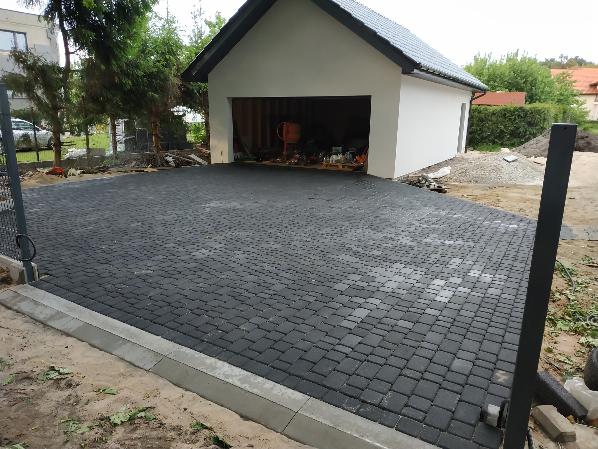 Nowoczesna, czarna kostka brukowa ułożona przed garażem z szarą podmurówką. Widoczne elementy ogrodzenia i przygotowany teren pod dalsze prace.