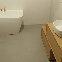 Instalco - Nowoczesna łazienka z białą wolnostojącą wanną, bidetem i umywalką nablatową na drewnianej szafce, armatura w kolorze złotym, szare płytki podłogowe.