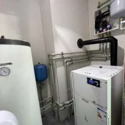 Węzeł cieplny w kotłowni: biały piec gazowy Wentor, zbiornik Kospel z manometrem, niebieski zbiornik wyrównawczy, liczne rury z mocowaniami, czarny komin, skrzynka elektryczna z manometrami...