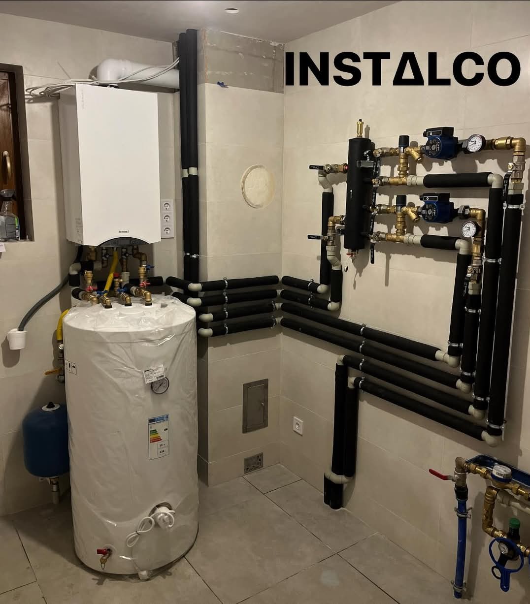 Kotłownia w trakcie instalacji: nowy biały zasobnik ciepłej wody owinięty folią, podłączony do kotła gazowego, z widocznymi izolowanymi rurami i elementami armatury. Na ścianie widoczne logo firmy...