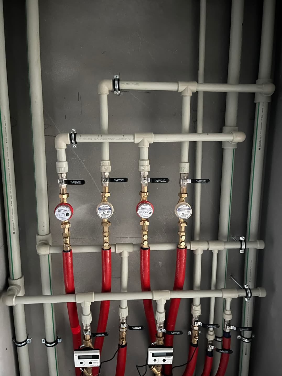 Szereg czterech wodomierzy z zaworami i czerwonymi rurami w izolacji, zamontowanych na białych rurach z tworzywa w szarej wnęce.