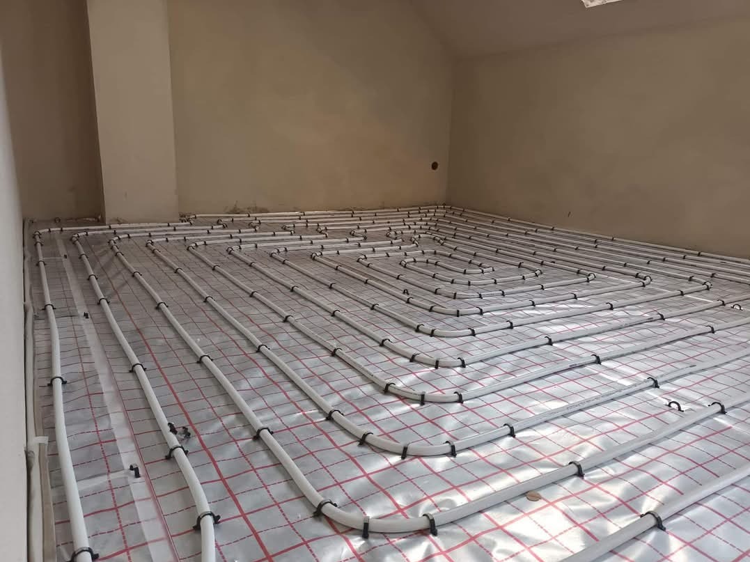 Białe rury ogrzewania podłogowego ułożone spiralnie na folii z czerwoną kratką w surowym pomieszczeniu, widoczne czarne uchwyty mocujące.
