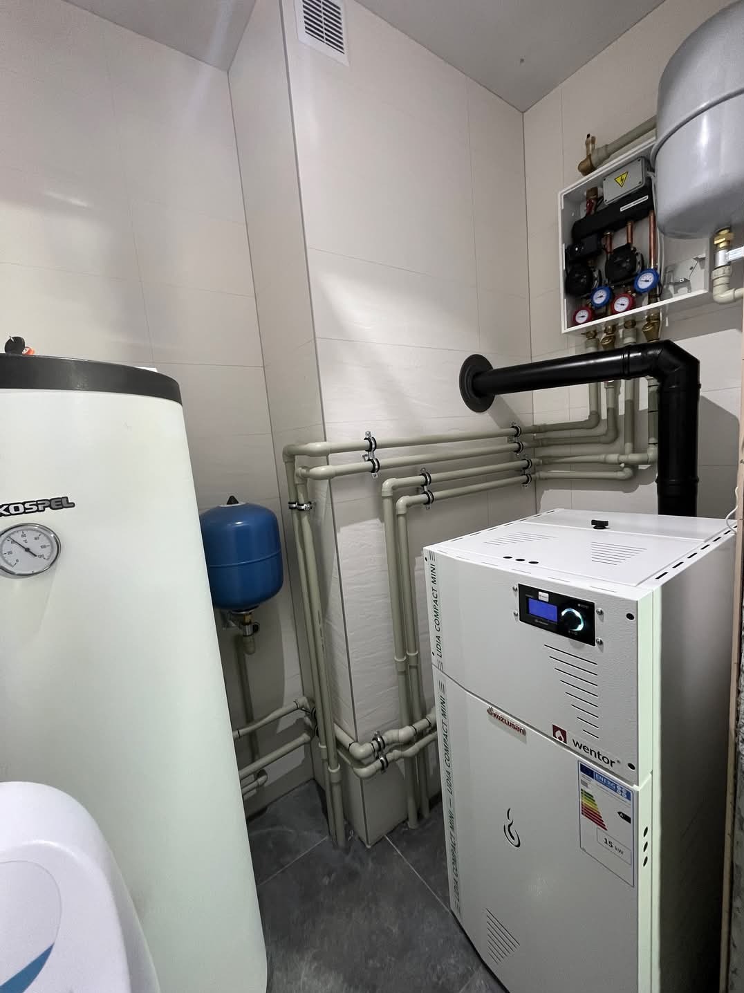 Węzeł cieplny w kotłowni: biały piec gazowy Wentor, zbiornik Kospel z manometrem, niebieski zbiornik wyrównawczy, liczne rury z mocowaniami, czarny komin, skrzynka elektryczna z manometrami...