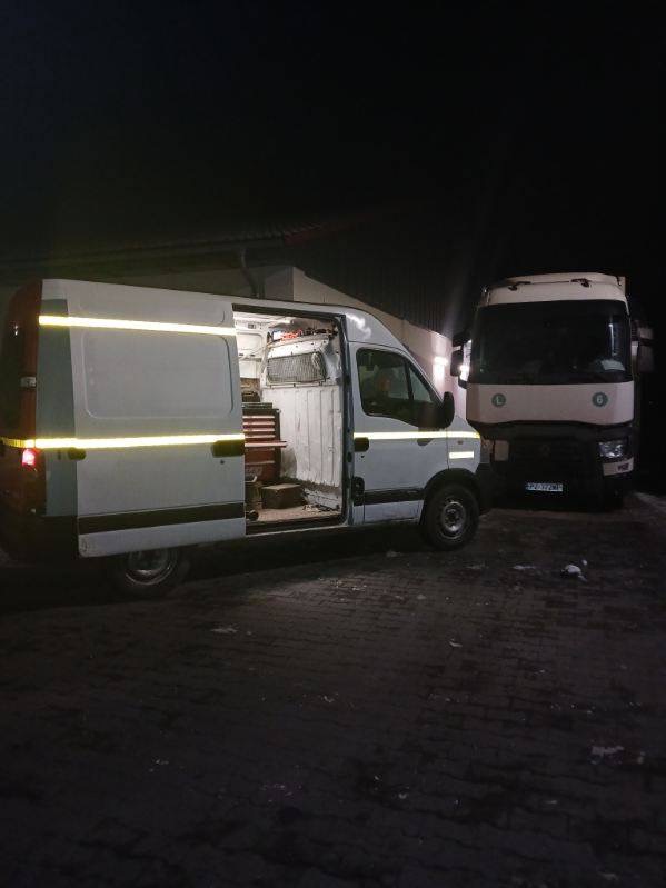 Biały samochód dostawczy z otwartymi drzwiami bocznymi, wewnątrz widoczny zestaw narzędzi; obok zaparkowany duży samochód ciężarowy, nocne oświetlenie.