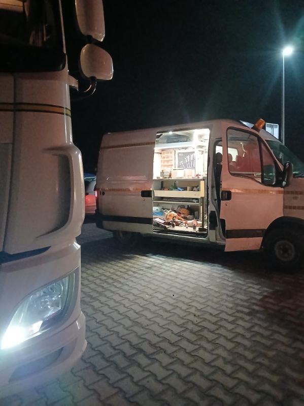 Mobilny warsztat: biały van z otwartymi drzwiami, wewnątrz narzędzia i oświetlenie. Obok zaparkowany biały samochód ciężarowy. Usługa na drodze.