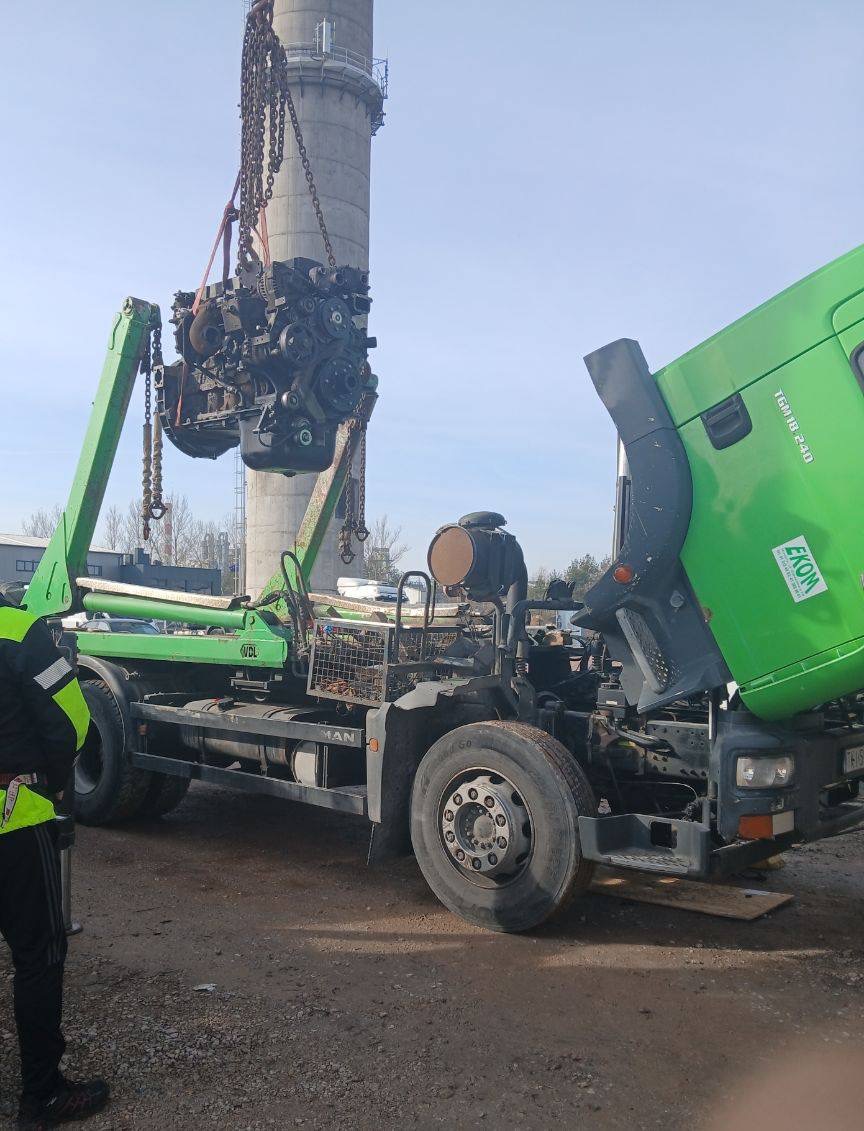 Zielona ciężarówka MAN z podniesioną kabiną, silnik zawieszony na łańcuchach, w tle komin. Mechanik w kamizelce odblaskowej obserwuje proces.