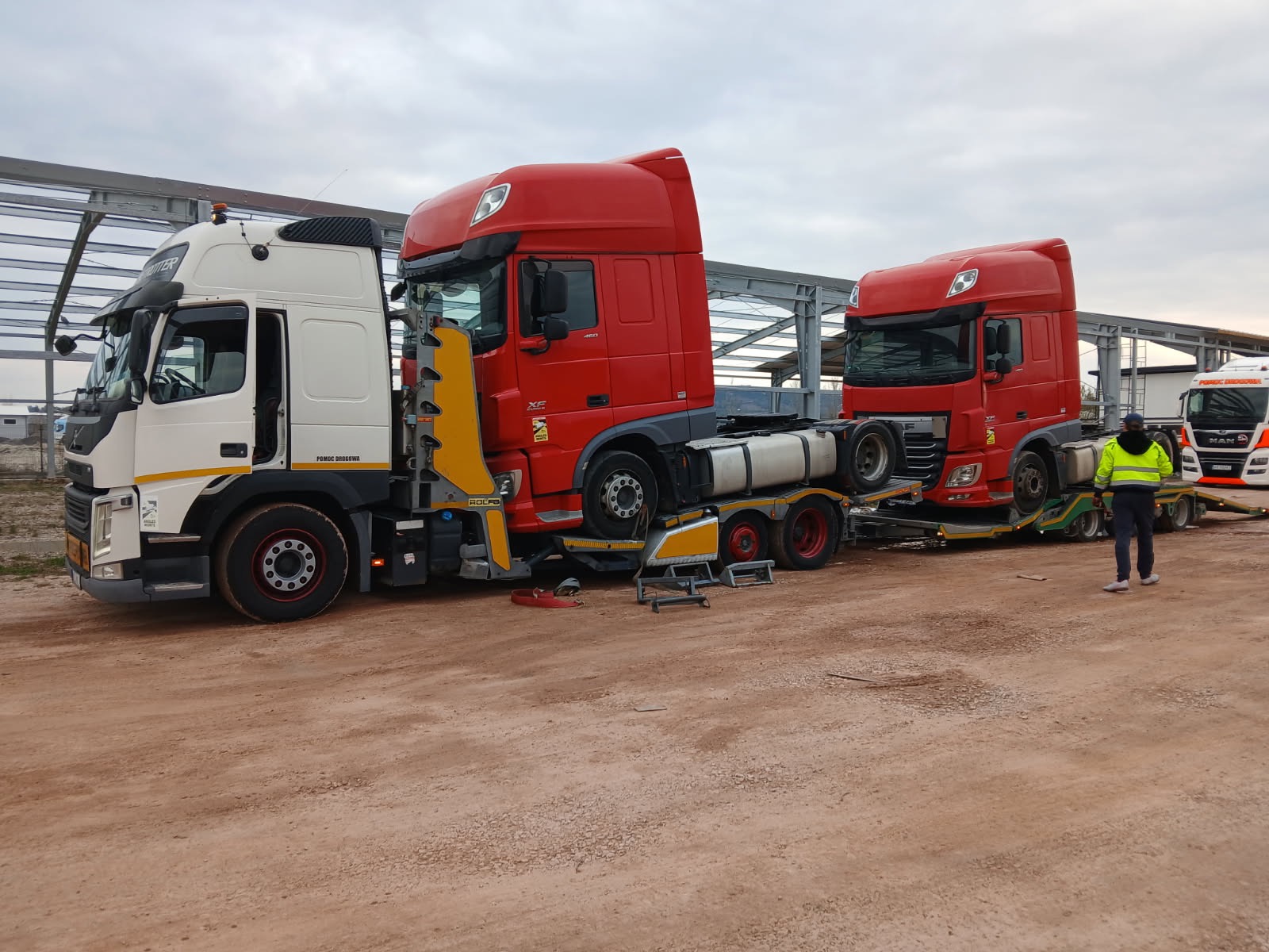 Ciężarówka pomocy drogowej z dwoma czerwonymi ciągnikami siodłowymi na lawecie, pracownik w odblaskowej kamizelce, teren budowy w tle.