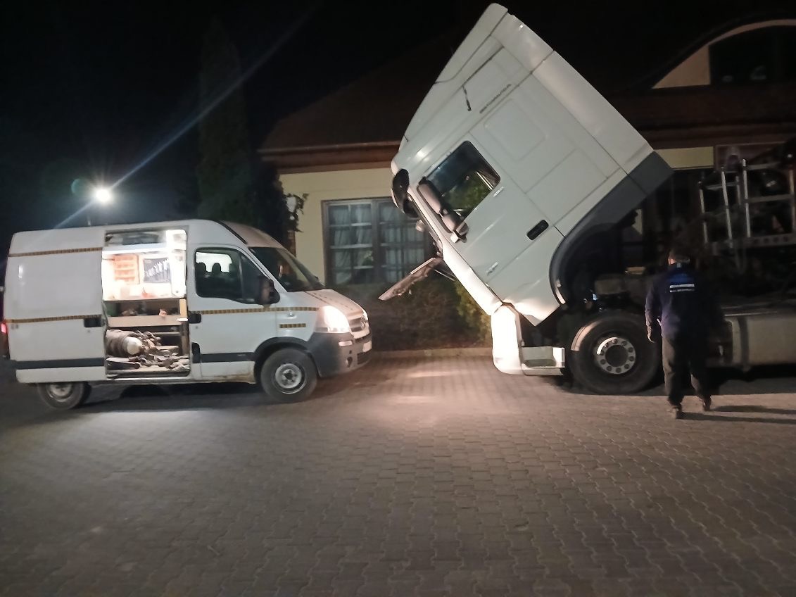 Awaria ciężarówki naprawiana nocą przez mobilny warsztat. Otwarty van serwisowy obok unieruchomionego pojazdu. Mechanik w ciemnym ubraniu przy ciężarówce.