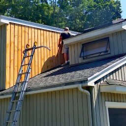 HANDYMAN - Pracownik stojący na dachu domu jednorodzinnego z drabina opartą o ścianę z surowego drewna, prawdopodobnie podczas montażu lub naprawy elewacji.