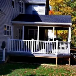 Zadaszony taras z białymi balustradami przylegający do domu z szarym sidingiem, widok na jesienne drzewa w tle.