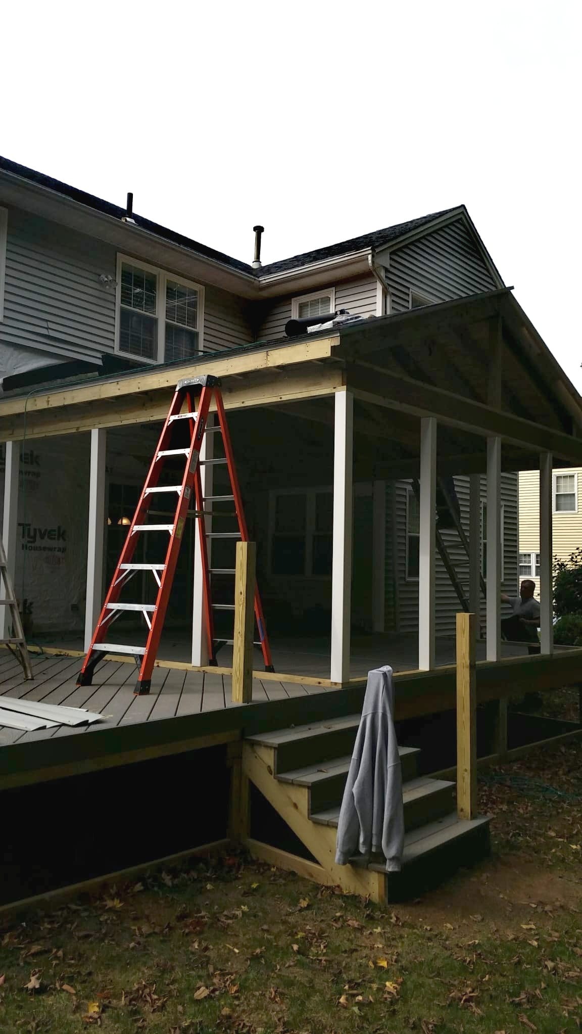 Budowa zadaszonego tarasu z drewnianą konstrukcją i pomarańczową drabiną, widoczny fragment domu z sidingiem i pracownik w tle.