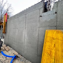 NAPOL SPÓŁKA Z OGRANICZONĄ ODPOWIEDZIALNOŚCIĄ - Świeżo wylana, segmentowa ściana fundamentowa z widocznym zbrojeniem u góry, szalunkami i niebieskim przewodem na ziemi. W tle widać drzewa i żuraw budowlany.
