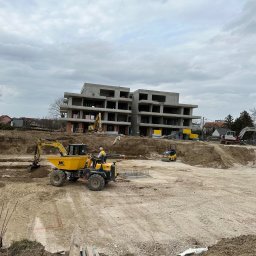 NAPOL SPÓŁKA Z OGRANICZONĄ ODPOWIEDZIALNOŚCIĄ - Budowa wielopiętrowego budynku w stanie surowym otwartym. Na pierwszym planie żółta wywrotka i minikoparka. W tle widoczny teren budowy z maszynami.