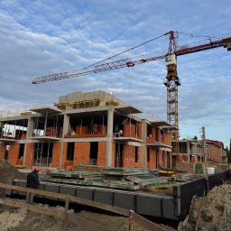 NAPOL SPÓŁKA Z OGRANICZONĄ ODPOWIEDZIALNOŚCIĄ - Wznoszenie dwukondygnacyjnego budynku z czerwonej cegły. Widoczny żuraw budowlany, pracownicy w kaskach i zabezpieczone fundamenty. Niebo z chmurami w tle.