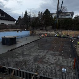 NAPOL SPÓŁKA Z OGRANICZONĄ ODPOWIEDZIALNOŚCIĄ - Wylewanie betonu na zbrojenie fundamentów domu jednorodzinnego. Widoczna pompa do betonu, pracownicy w odblaskowych ubraniach i szalunki. Pochmurne niebo w tle.