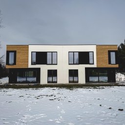 NAPOL SPÓŁKA Z OGRANICZONĄ ODPOWIEDZIALNOŚCIĄ - Nowoczesny, dwukondygnacyjny dom z elementami drewnianej elewacji i dużymi oknami, częściowo pokryty śniegiem, na tle pochmurnego nieba.