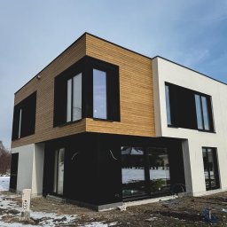NAPOL SPÓŁKA Z OGRANICZONĄ ODPOWIEDZIALNOŚCIĄ - Nowoczesny dom z drewnianą elewacją i czarnymi ramami okiennymi, częściowo otynkowany na biało. Widoczne ślady budowy i resztki śniegu na trawie.