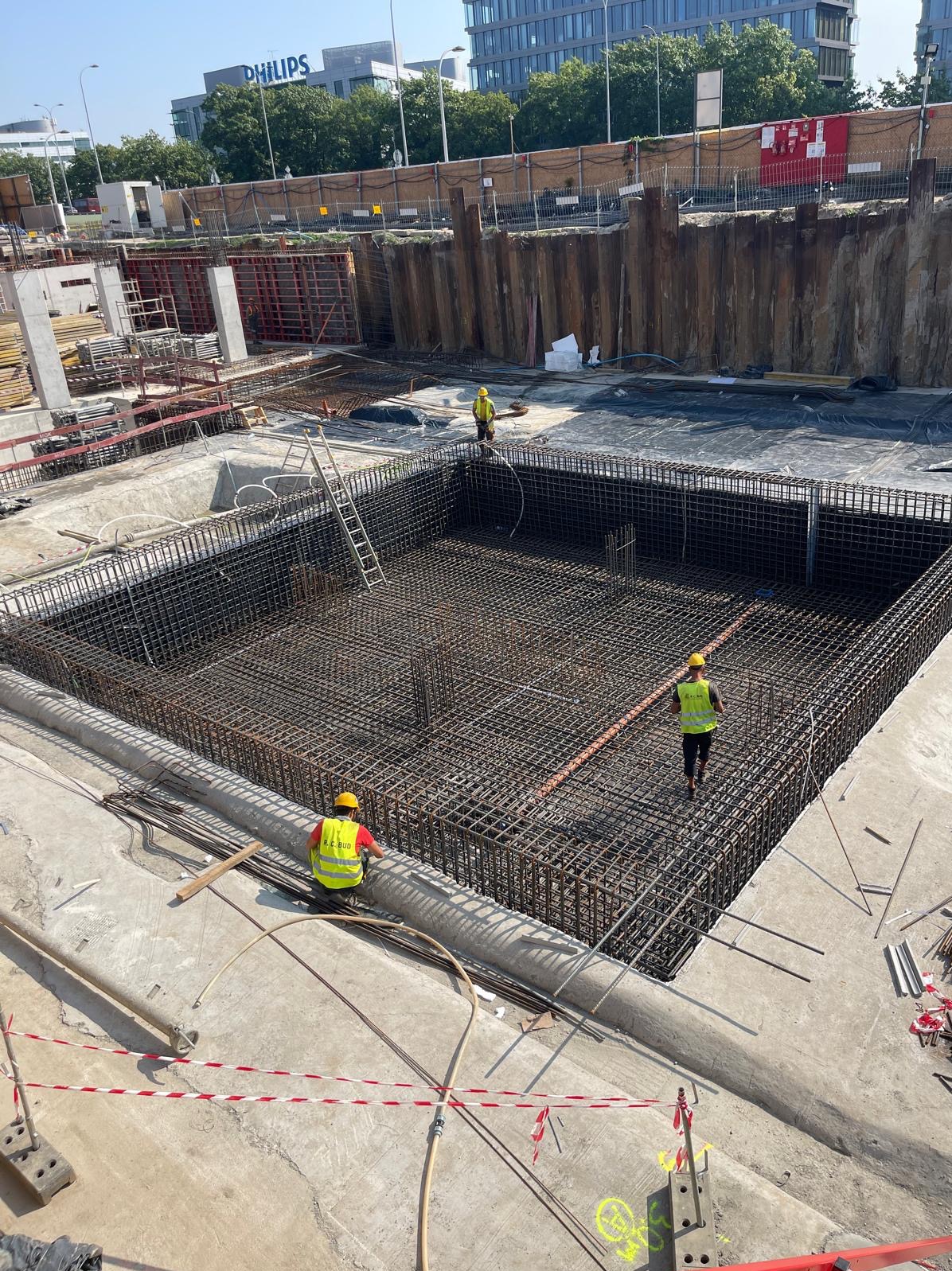 Zbrojenie fundamentu w wykopie, widoczne stalowe pręty, szalunki, pracownicy w kaskach i kamizelkach odblaskowych, tło z budynkami biurowymi.