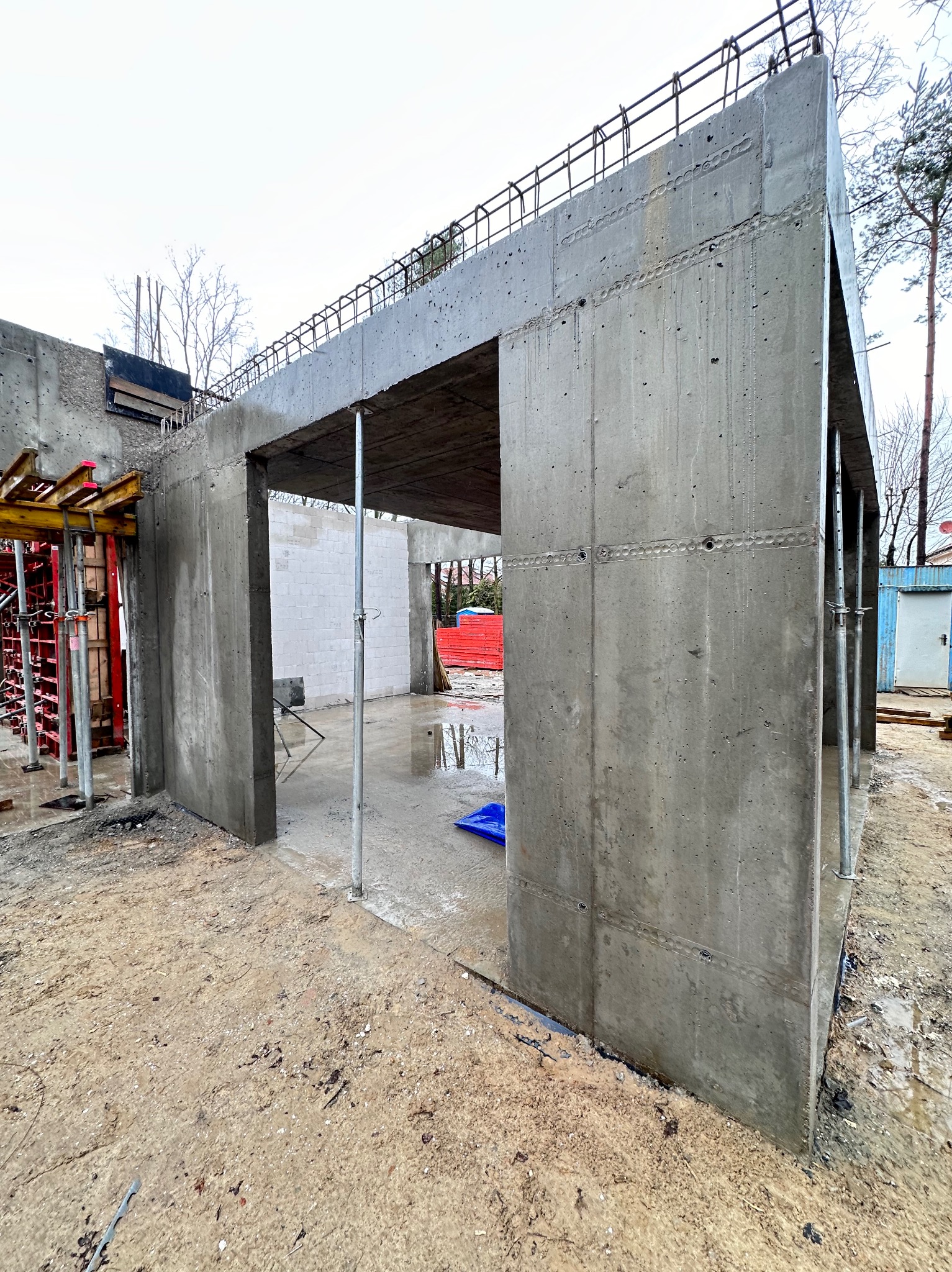 Surowa konstrukcja żelbetowa w trakcie budowy. Widoczne szalunki, zbrojenia i podpory. W tle teren budowy z elementami czerwonych materiałów budowlanych.