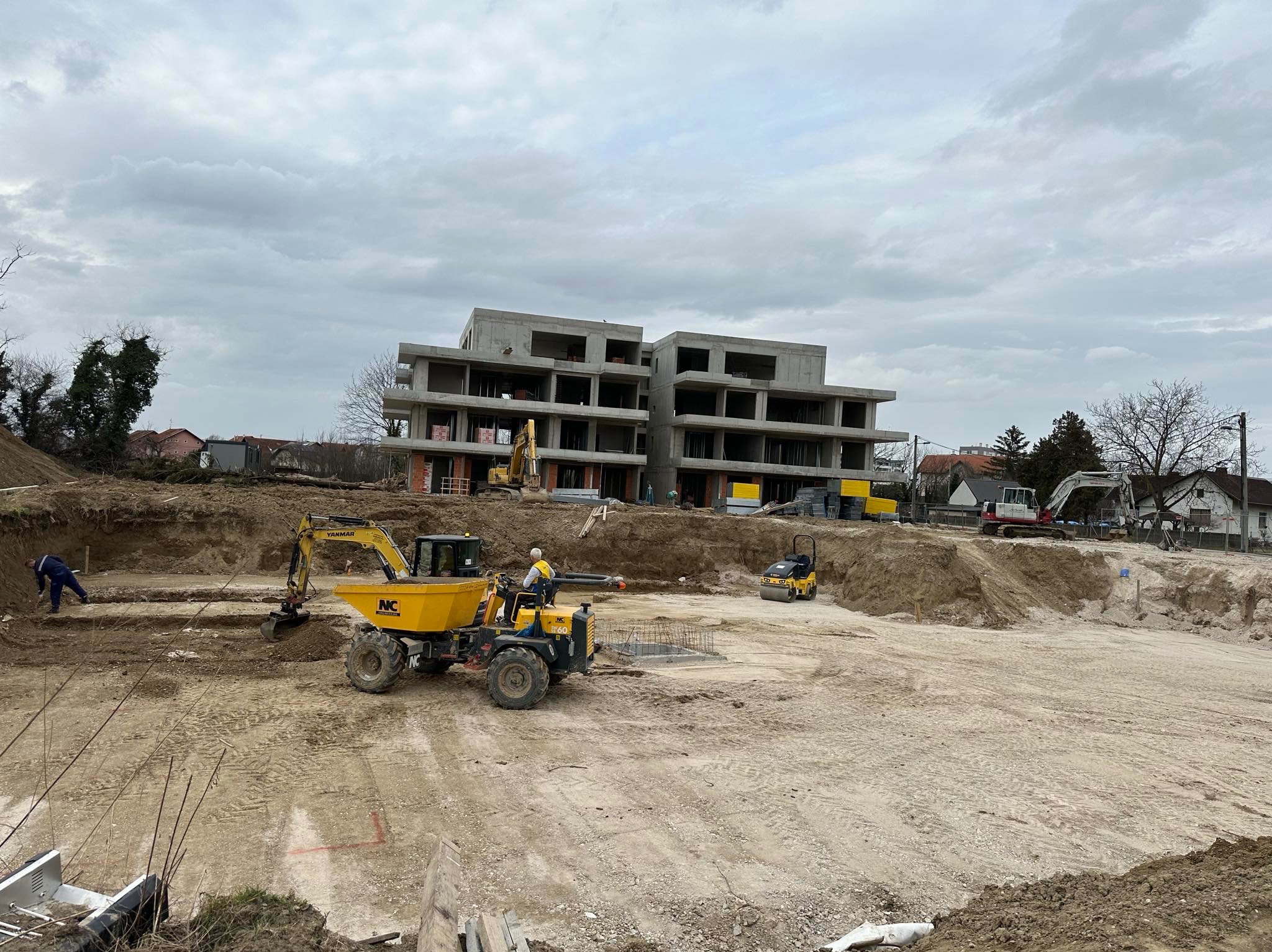 Budowa wielopiętrowego budynku w stanie surowym otwartym. Na pierwszym planie żółta wywrotka i minikoparka. W tle widoczny teren budowy z maszynami.