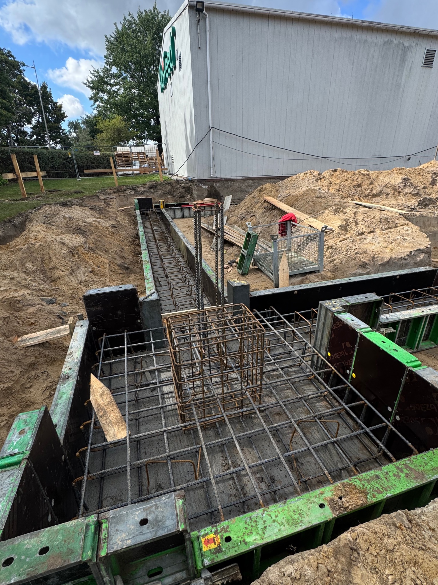 Zbrojenie fundamentów w szalunkach z zielonymi elementami, widok z góry. Stalowe pręty tworzą siatkę i klatkę, przygotowane do zalania betonem. Prace ziemne w tle.