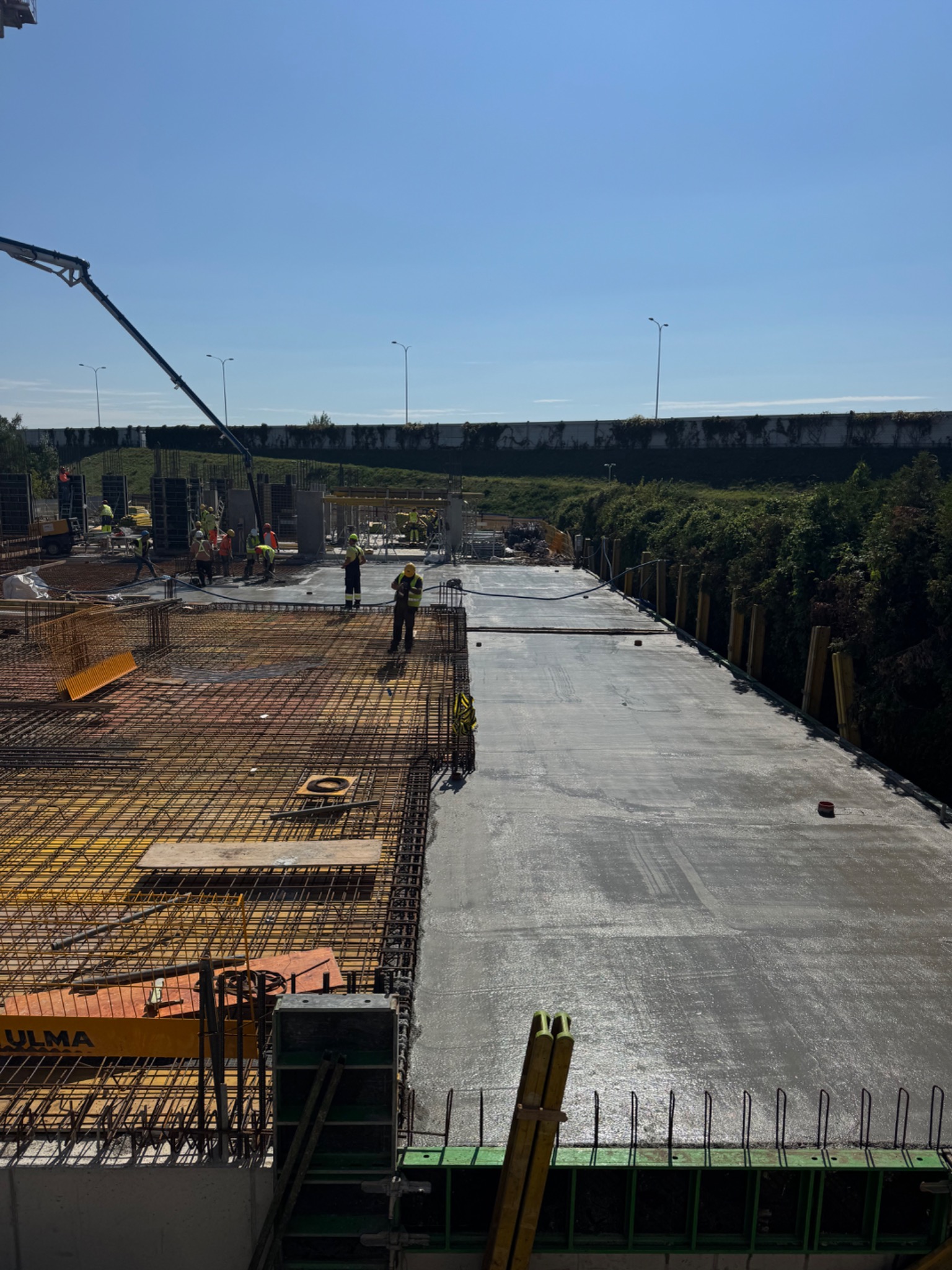 Wysoki kąt: Zbrojenie i wylewanie betonu na budowie. Widoczni pracownicy w kaskach i kamizelkach. Część powierzchni już zalana, część przygotowana zbrojeniem.