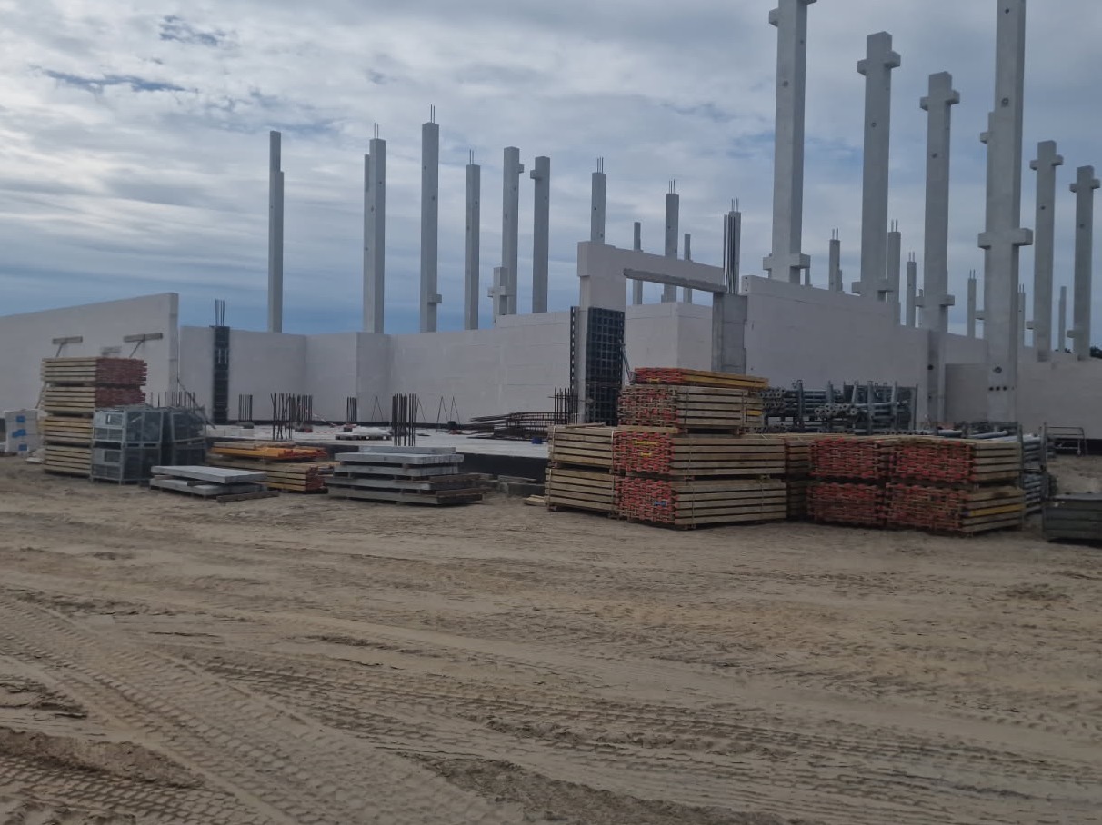 Wznoszenie budynku żelbetowego: widoczne betonowe słupy, ściany i przygotowane szalunki na placu budowy. Surowy stan, pochmurne niebo w tle.